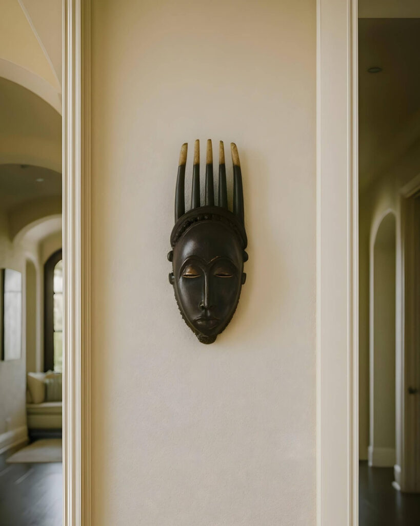 A dark wooden African Baule mask with closed eyes and decorative headdress is mounted on a beige wall in a hallway, creating a serene and cultural ambiance.