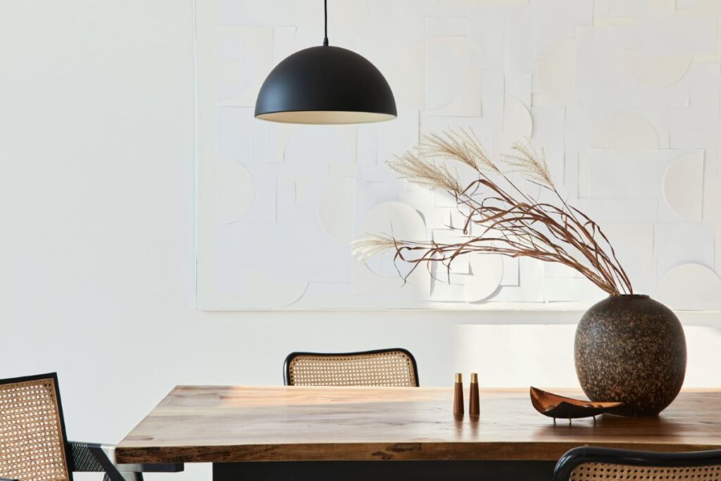 Close up on a wooden dining table with organic decor on it. Clean lines and modern aesthetic, a round clay vase, a black metal lamp and contemporary chairs 