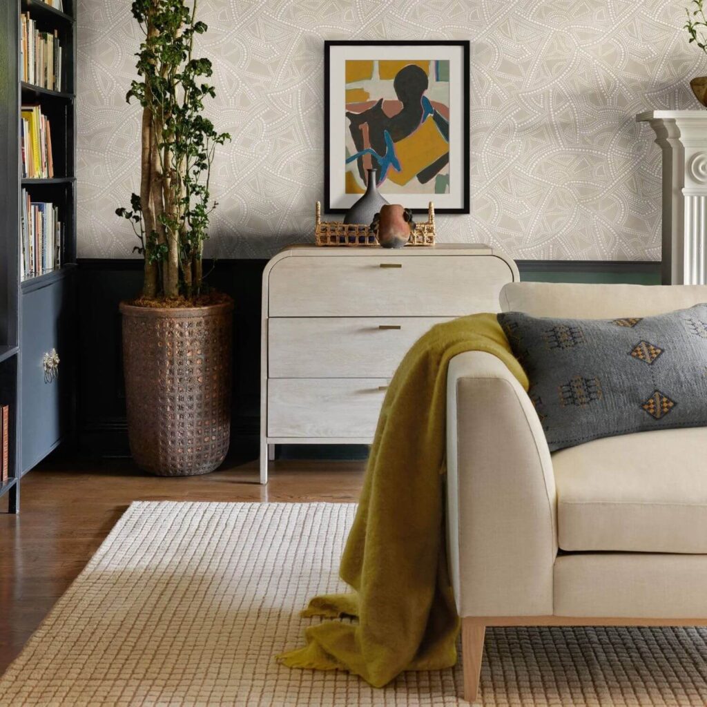 Cozy living room area with an off white african-inspired wallpaper, an off-white rug, cabinet and sofa, a mustard throw blanket with a blue throw pillow, a plant, some art and books in a Library