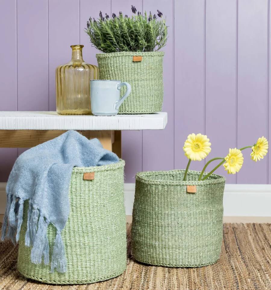 Three sage green sisal baskets. The larger one has a baby blue scarf in it, the medium basket has sunflower in it and the smaller one has a bushy plant in it.