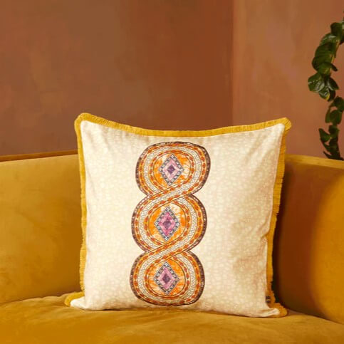 Gifts for Interior Design lovers - A beige cushion with a bright orange African pattern the middle that has a circular braid-like shape with accents of pink and purple. The cushion also has a yellow border