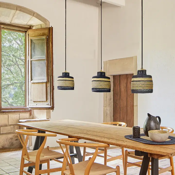 Cylindric-shaped unique pendant lights in natural and navy colors, woven in Ghana with durable grass and hanged over a natural wooden dining table with wooden chairs