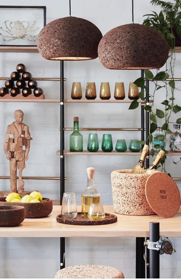 Two brown cork pendant lights installed over an elegant bar stocked with liquor and champagne. Cork bowls, a cork platter and a cork bucket are on the bar. Behind the bar are shelves with glasses in dark yellow and green shade.