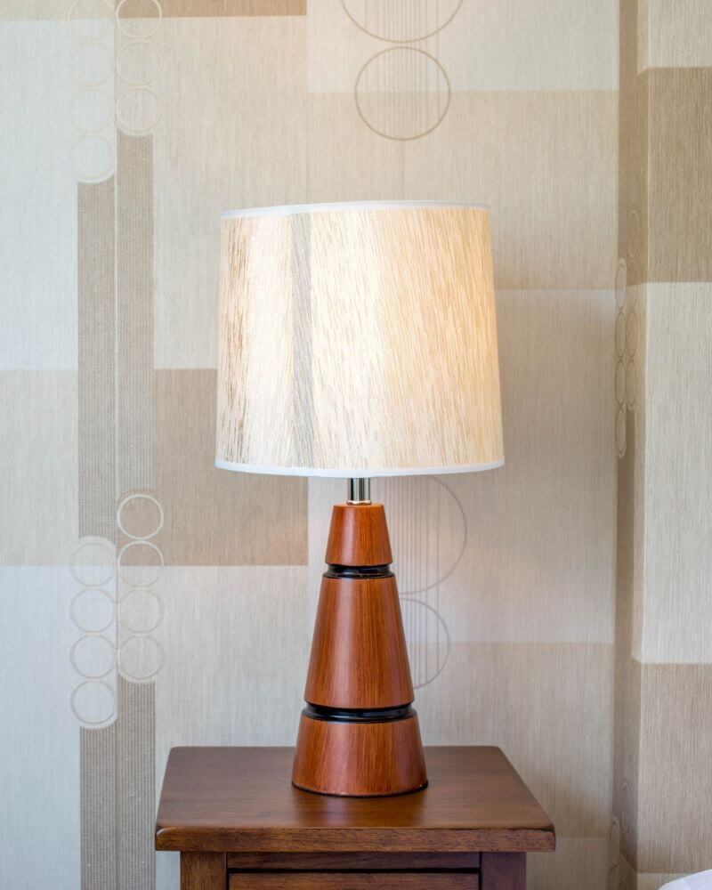 Table lamp with a conical wooden base and a beige lamp shade on a bedside table in front of a wall with a beige wallpaper