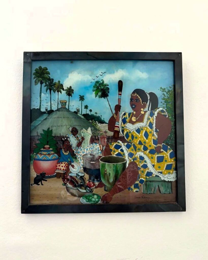 Senegalese underglass painting depicting a mundane village scene: woman dressed traditionally but elegantly cooking with a pestle and mortar. Man reading the newspaper in the background and young girl washing the dishes.