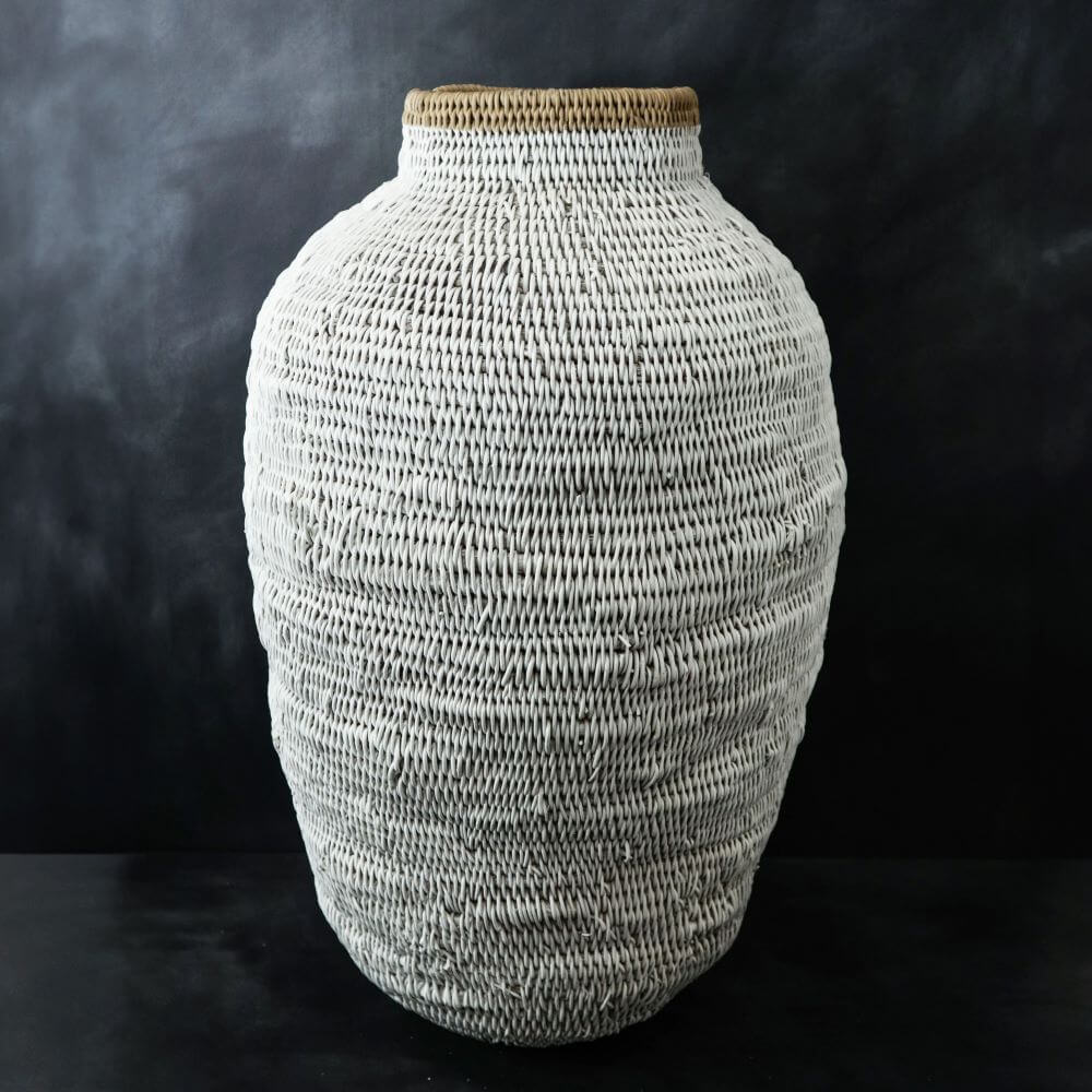 The captivating image features a large, handcrafted buhera vase basket, exuding an earthy charm with its intricate woven texture. The vase is an organic, rounded shape, widest at the middle and tapering gently towards the bottom, then narrowing as it rises to a smaller opening at the top. The surface of the vase is entirely covered in an off-white woven pattern, providing a rustic yet elegant aesthetic. The pattern is constructed from thin, fibrous material that's carefully intertwined to create a visually appealing texture, with the weave adding depth and giving the vase dimension. Crowning the top of the vase is a rim made of a natural, light brown material, which further accentuates the pot’s handmade design. The rim's coarse texture contrasts beautifully with the smooth, woven body, adding complexity to the design. The backdrop behind the vase is a dark, almost black, color which serves as a stark canvas. The contrast between the dark background and the light vase makes the details of the weave stand out more. Situated on a dark surface as well, the vase has a grounding presence, making it the focal point of the image.