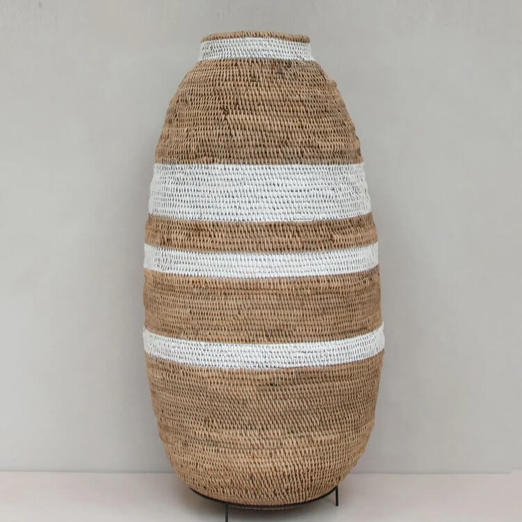 The photo features a tall, large, vase-like buhera basket standing vertically. The basket is made of natural brown material and is decorated with horizontal stripes. The predominant color of the basket is a light tan or beige, which looks like a woven material with a visible texture characteristic of natural fibers. The basket is embellished with several wide, white-painted stripes that run horizontally around the circumference, contrasting with the natural brown of the basket. The overall shape of the basket is slightly bulbous, resembling a large egg with a flattened top. The woven texture is consistent throughout the body of the basket, suggesting handmade craftsmanship. The basket sits on a minimal, wrought-iron stand, providing a slight lift from the surface it's placed on. The background of the photo is a plain, light-colored wall, which helps to emphasize the basket as the primary subject. The lighting appears to be soft and even, which highlights the textures and colors of the basket without harsh shadows.