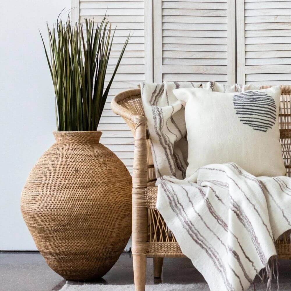 Large buhera basket holding tall green grass placed next to a woven chair from Malawi draped with a cozy cotton throw and beautiful throw pillow with a wool cover