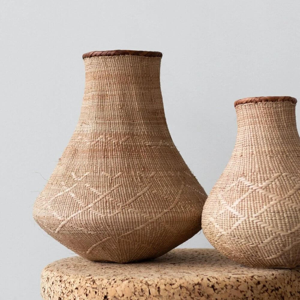 The image shows two vase-shaped woven gourd African baskets against a plain white backdrop. The basket stands upright and is the central focus of the composition. The basket is predominantly light brown or tan in color, with a distinct woven texture that is tight and intricate. It has a wide, bulbous base that gradually tapers inward as it rises, forming a narrow neck at the top. The rim of the basket is finished with a brown binding or trim. The basket appears to be handmade, with slight variations in the weaving pattern adding to its rustic charm. The light catches the woven surface, highlighting the texture and subtle color variations within the natural material. It has a large base compared to the narrow top, and it is of an equal color throughout.