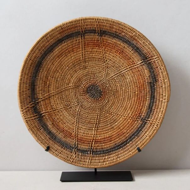 A circular woven Makenge basket is displayed on a black stand against a neutral background. The basket, likely made of natural fibers, exhibits a complex spiral weave pattern emanating from a dark-colored central point. This design gives the impression of radiating lines, similar to the rings of a tree trunk. A distinct circular band of dark color surrounds the basket's center, adding depth and contrast to the design. The primary color of the basket alternates between warm tan and orange hues, suggesting the use of various natural materials and dyes in its construction. The black metal stand features a rectangular base and a minimalist design, drawing attention to the basket itself. The backdrop is plain and off-white, which does not distract from the intricate details of the basketry. The lighting is soft and evenly distributed, minimizing harsh shadows and highlighting the texture of the woven material. The overall composition emphasizes the craftsmanship of the basket and its role as a decorative object.