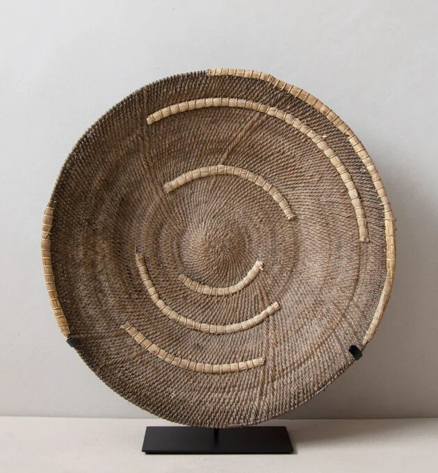 The image features a large, circular woven Makenge basket displayed on a black metal stand against an off-white background. The basket is the focal point, with its intricate spiral pattern and texture creating a sense of depth. The basket appears to be made of natural fibers, likely straw or a similar material. Its color is a mix of earthy tones, ranging from light beige to a darker brown, giving it a rustic and organic feel. The weaving is tight and uniform, except for the occasional loop of what seems to be straw around the outside, indicating that some straw fibers were formed. The basket has a shallow, bowl-like shape, further emphasizing its artisanal craftsmanship. Around it, there are straw pieces that create arcs around the basket, that get smaller and smaller to the basket makes in a spiral. The black metal stand beneath the basket provides a stark contrast. The stand blends into the background. The off-white background creates a sense of minimalism, directing the viewer's attention solely to the basket and its stand. The image is well-lit, highlighting the textures and patterns of the basket while maintaining a soft, natural ambiance. The absence of other distracting elements ensures that the focus remains on the basket as a decorative object.