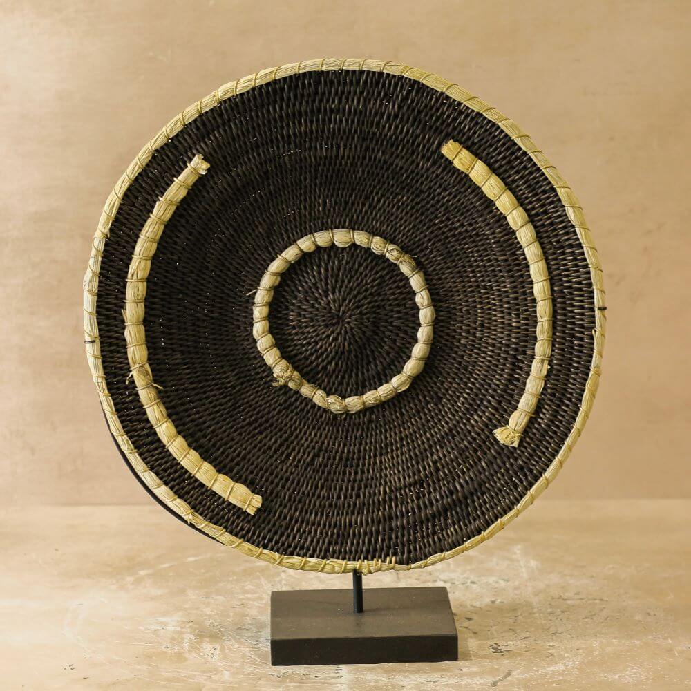 A close-up shot showcases a handcrafted, circular woven wall Makenge basket displayed on a simple stand. The basket-like piece is predominantly dark brown, exhibiting an intricate woven pattern throughout its surface. The decoration features two prominent embellishments: a circular ring at its center and a larger, semi-circular band that traces a portion of the outer edge. Both of these adornments are crafted from a lighter, straw-colored material, providing a striking contrast to the dark brown backdrop. The straw elements appear to be sections of tightly bound grass or similar natural fiber, lending a rustic, organic feel to the piece. The decoration is secured to a minimalist stand, which consists of a thin, vertical metal rod affixed to a square, dark-colored base. The base sits on a light-colored surface with subtle marbled patterns, suggesting a stone or composite countertop. In the background, the wall is painted in a neutral, beige tone, providing a soft, unobtrusive backdrop that allows the decorative piece to stand out. The lighting in the shot is diffused, casting gentle shadows and highlighting the textures of the woven material. The composition of the image focuses sharply on the wall decoration, minimizing distractions and emphasizing the craftsmanship and design of the piece.