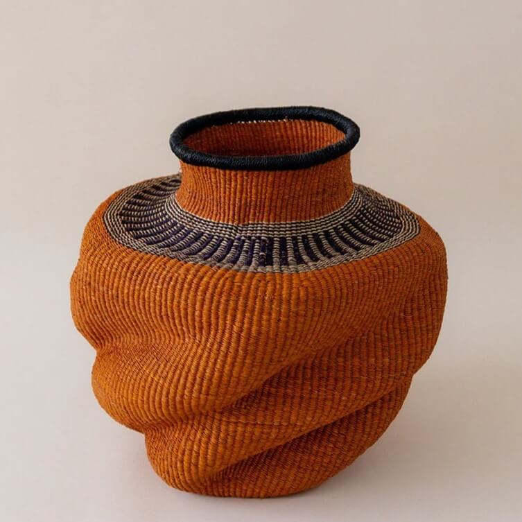 The image shows a hand-woven vase basket, likely from natural fibers or reeds, in a warm, earthy palette. The vase features a distinctive design with a stout body that appears to be twisted or spiraled as the weaving progresses upwards. The overall color is predominantly a rich terra cotta or reddish-brown, giving it a natural and rustic appearance. At the top of the vase, there’s a defined neck which is also woven, leading to a round opening. The rim of the opening is finished with a black material, adding a touch of contrast and definition to the top edge. Just below the neck, there's a band of darker-colored weaving, possibly darker shades of brown or even navy, patterned in neatly arranged horizontal lines. This contrast highlights the cylindrical form of the neck before it flares out to the body of the vase. The texture of the weaving is evident, showcasing the individual strands or reeds that have been meticulously intertwined. This gives the vase a tactile quality, suggesting the time and skill that went into its creation. The vase is set against a neutral backdrop, which could be a wall or surface, allowing the object to be the sole focus. The lighting seems soft and even, minimizing harsh shadows and highlighting the vase’s three-dimensional form and texture. Overall, the piece has an organic, artisanal aesthetic, likely intended as a decorative item that would complement either a modern or more bohemian-style interior. The photograph itself is well-composed, providing a clear and detailed representation of the vase, emphasizing its unique design and craftsmanship.