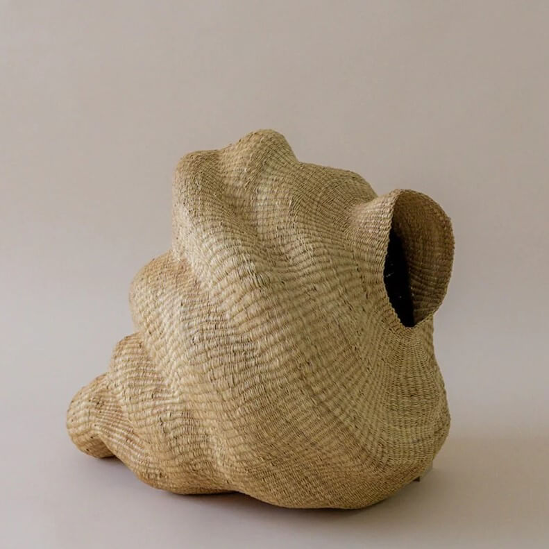 The image features a handcrafted woven basket with an abstract artistic design. The basket is made from natural fibers, showcasing a light beige hue, arranged in an intricate pattern. The weaving technique highlights thin, horizontally oriented fibers woven together. The basket has an organic, irregular shape, with a form that appears undulating. It is widest at the base, then curves upwards, and then the the upper portion cascades downwards, culminating in a large, prominent opening that is seemingly the entry point. The basket sits on a plain white surface against a solid, neutral background. The lighting is soft and subdued, highlighting the textures and shapes of the woven material.