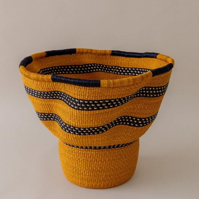 The image depicts a handcrafted woven basket in a warm mustard yellow hue, complemented by bands of dark blue and white dotted stripes. The basket is set against a neutral, light-colored background that casts a soft glow, showcasing the intricate texture of the weaving. The basket is constructed in two distinct sections. The upper portion flares outward from a narrow point, creating a vase-like shape that gently widens towards the top. This section features the mustard yellow base color interrupted by horizontal stripes of dark blue with white dotted patterns, adding a rhythmic visual texture. These stripes are evenly spaced, offering a repeating pattern that is both eye-catching and harmonious. The rim of the basket is reinforced with a band of the same dark blue, providing a clean, finished edge. Below this flared section, the basket tapers inward to form a cylindrical base. This base retains the mustard yellow color throughout, devoid of any striping. The transition between the flared top and the cylindrical base is smooth, indicative of skilled craftsmanship. The weaving technique appears consistent across the basket, maintaining an even texture and structure. The tight, uniform weaving highlights the natural fibers used, which suggests a sturdy and durable construction. The play of colors and the shape of the basket suggest traditional weaving techniques, likely from a culture known for its textile arts. The lighting accentuates the texture of the woven material, emphasizing the tactile quality of the basket. Overall, the image is a celebration of craftsmanship, showcasing a functional art piece with attention to detail and quality.