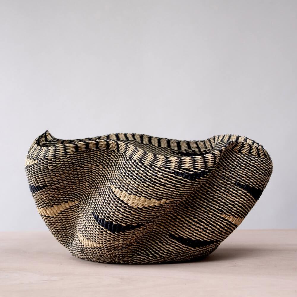 The image features a woven pakurigo wave basket with a distinctive, organically wavy silhouette made from natural fibers in a black and tan color scheme. The basket sits on a pale wooden surface against a plain white backdrop, emphasizing its unique shape and texture. The basket's construction is characterized by a tight weave, displaying alternating bands of black and tan that create a striped pattern. The colors appear to be derived from the natural hues of the fibers used. The top edge of the basket is undulating, giving it a soft, informal look. The organic shape gives the basket a sense of character. The basket sits directly on the wooden surface, its rounded base conforming to the plane. The texture of the fibers is palpable; the rough weave suggests the raw, natural materials. The plain white background further enhances the basket's presence, allowing the viewers to focus on the object. The pale wood provides a minimalist base, adding to the image's clean aesthetic.