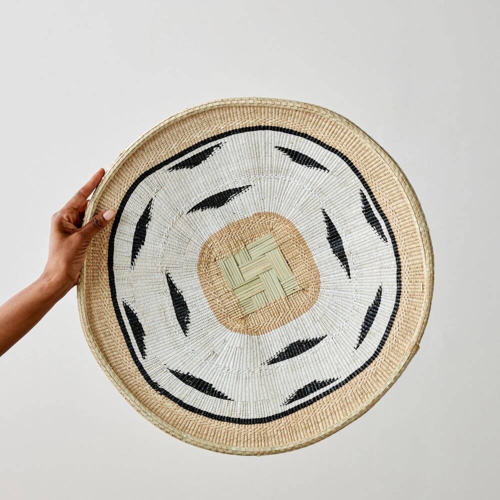 The image depicts a hand holding a decorative woven African wall basket with a square design in light green and light brown tones. Surrounding this square is a similar light brown band, followed by a white woven area with scattered black tear-shaped patterns that add a modern yet organic feel. The basket is edged with a continuous rim made of natural-colored woven material. The weaving around the rim is finer and more densely packed, providing a textural contrast to the patterns within. A person's hand with brown skin partially holds the left side of the basket, providing scale and context to the object. The background is a plain, light color, ensuring that the basket and hand are the focal points of the image. The lighting is soft and diffused, which highlights the textures and patterns of the woven materials without creating harsh shadows. The overall composition is balanced, with the basket's circular shape centered in the frame.