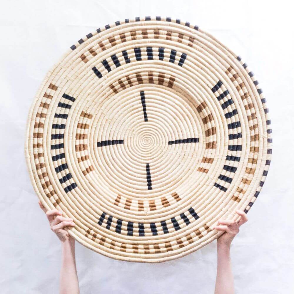 The photograph showcases a circular, intricately woven wall basket held aloft by two hands against a clean, white backdrop. The basket's construction is defined by concentric rings made from a light, straw-colored natural fiber, creating a spiral effect from its center to the edge. The basket's design incorporates dark brown and black accents interwoven into the straw base. These colors form geometric shapes: short, parallel lines are arranged in segments around the basket, creating a pattern interrupted by the straw color and other segments of brown. The innermost design features four thick, equally spaced black lines that extend from the center towards the edge, resembling the arms of a compass. Light from above illuminates the textured surface of the basket, casting subtle shadows within the weaves and highlighting the craftsmanship. The bottom portion of the frame shows the hands clasped tightly around the bottom edge of the basket. The overall image evokes a sense of artisanal creation and careful attention to detail, emphasizing the basket's visual appeal and tactile quality.