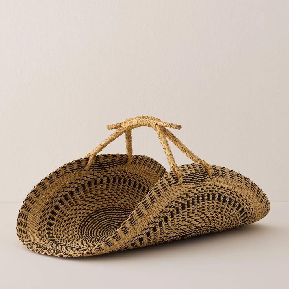 The photograph features a woven, tan-colored basket with black geometric patterns, set against a stark white background. The basket has a structural design, with a semi-circular shape and two handles that come together to form one single handle. The African baskets have a textured design, created by the pattern of light and black material. The edges of the baskets are defined with a band of light brown, and the semi-circular shape of the baskets is clearly visible. The weaving of the baskets is visible, with lines and textures creating an intricate design. The black geometric patterns add contrast to the otherwise monotone tan color of the baskets. The lighting is soft and even, with no harsh shadows. The texture of the woven material is visible, adding depth and interest to the image. The overall tone of the image is simple and clean, with the focus on the baskets and their unique design.