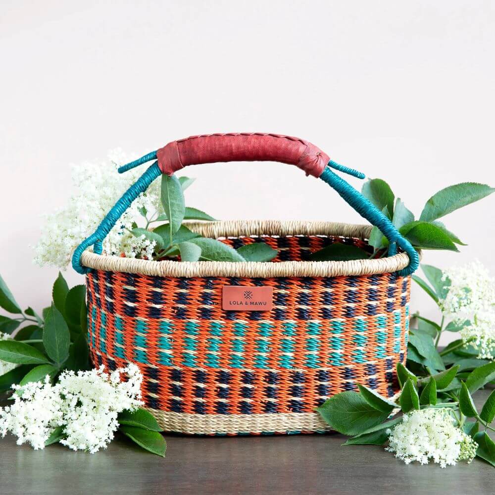 The image showcases a woven African basket, exuding a blend of rustic charm and artisanal craftsmanship. The basket is primarily orange with interspersed patterns of blue-green and navy-blue that form textured rows. The bottom of the basket and the top rim are woven from a neutral, straw-like color, providing a natural border. Attached to the basket are handles wrapped in a vibrant teal color, leading up to a textured leather grip that is a deep red-brown hue. The leather adds a touch of sophistication and durability to the basket. A small, rectangular label is affixed to the center of the basket and showcases the brand name "LOLA & MAWU," possibly a maker or designer's mark. Strategically placed around the basket are bountiful sprigs of white flowers with lush green leaves, giving the impression that the basket is overflowing with flora. Some of the foliage is inside the basket, while the rest frames it from the sides and bottom. The background is a soft, light peach color, which accentuates the basket and greenery. The photograph appears to be taken in a well-lit environment, capturing the textures and colors in detail. The presentation suggests that the basket may be a decorative item or a storage solution being marketed for its aesthetic and practical qualities.