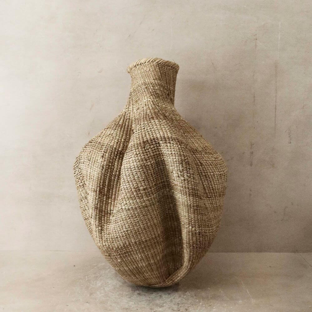 The image shows a tall, woven Ilala gourd basket in a neutral, earth-tone color. The basket stands upright on a slightly mottled gray surface, likely a concrete or stone floor. Behind the basket is a wall of a similar neutral shade, providing a backdrop that highlights the texture and form of the basket. The basket itself has a unique, bulbous shape, wider at the bottom and tapering into a narrow neck towards the top. The woven material appears to be some kind of natural fiber, such as straw or reeds, and it exhibits a dense, even weave that creates a corrugated texture across the surface. The weaving lines are oriented vertically for the most part, following the curves of the basket, emphasizing its organic shape. At the very top, the basket finishes with a tightly bound rim, slightly darker in color, creating a subtle contrast. The muted palette and the rough texture of the weaving give the basket a rustic, artisanal feel, making it a striking decorative object.