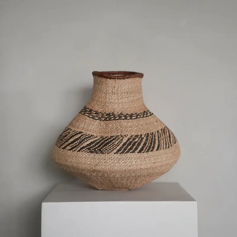 A woven gourd basket is placed on a square, white pedestal. The basket is light brown and has a medium-size opening, with the space surrounding the inner rim lined with darker brown braiding. The top of the basket is cylindrical for about 3.5 inches, and then the body of the basket bows out into its widest part. The base of the basket is flat so that it can sit on the pedestal. There are two distinct sets of patterns. At its widest, there is a set of 3 black stripes created by small sets of diagonal lines. Near the top of the basket there is a set of zig-zagging lines. The background consists of a plain wall with the top of the basket reaching about 55% of the height of the frame. The image is well-lit; there are no major shadows except for a slight gradation of darkness on the base of the pedestal.