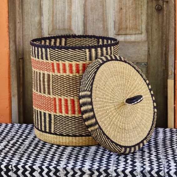 The photograph features a large, handwoven laundry basket with a lid, set against a rustic background. The basket is cylindrical in shape, with a complex pattern of horizontal and vertical stripes in natural straw yellow, black, and red hues. The lid, which leans against the side of the basket, has a similar woven design, with a black and natural straw yellow pattern radiating from the center. A small, dark handle is visible on the top of the lid. The basket sits on a black and white chevron-patterned surface, possibly a rug or tablecloth. The background consists of a weathered wooden door with vertical panels and a faded orange wall on the left side of the frame. This door is situated behind the basket, adding depth to the composition. The lighting is soft and diffused, highlighting the texture of the woven materials and the aged appearance of the wooden door. The overall scene has a warm, inviting feel, suggesting a handcrafted or artisanal product on display.