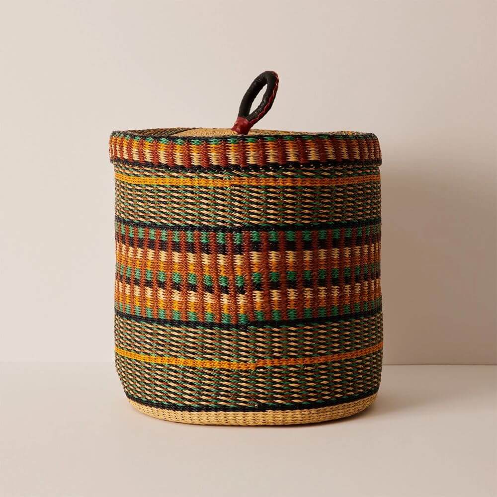 The image shows a cylindrical woven basket. The basket is predominantly green and brown, with horizontal stripes of black and yellow. Rectangles of black and golden brown are also woven into the basket's design. The bottom of the basket is light brown. On top, there's a flat lid made in the same basket-weaving style, and atop the lid, there is a handle made of black material with red accents. The background is a plain, neutral color, which allows the details of the intricate basket to become the focal point.