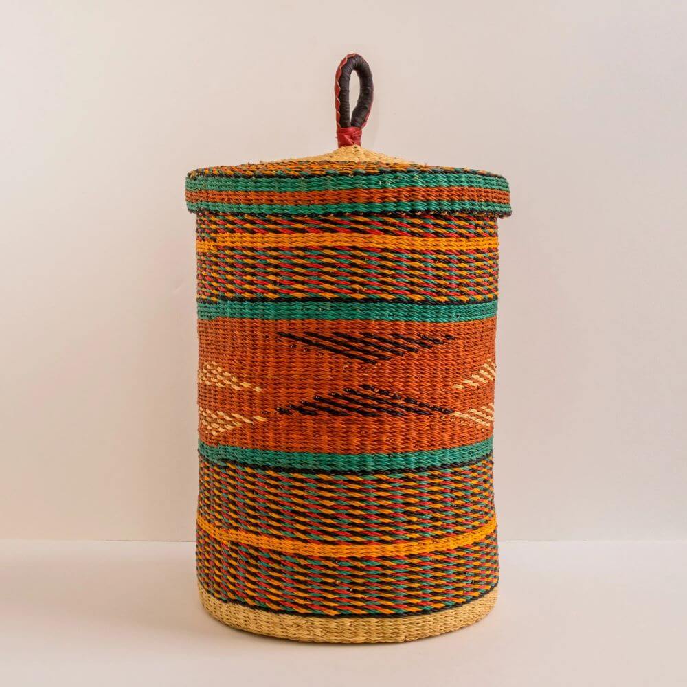 The image shows a cylindrical woven laundry basket with a lid, set against a simple, light-colored background. The basket is made using woven materials in a variety of colors that give it a striped or multi-patterned effect. The top of the basket is covered with a lid that has a loop handle at the center. The handle has a red band at the joint of the loop with the lid. The lid fits snugly on the basket's top, and its edges also feature the woven pattern, matching the primary color scheme. The sides of the basket prominently display horizontal stripes and geometric patterns. There are bands of color alternating between orange, yellow, green, and thin black lines. The orange sections feature patterns woven into the basket, creating diagonal lines and chevron designs. The base of the basket has a different weave, giving it a golden, natural material look. The weave texture is consistent throughout the body of the basket, which hints at the handmade nature of the item. The background is a flat, off-white color that does not distract from the subject. The lighting is soft and evenly distributed, which highlights the colors and intricate texture of the basket. The basket appears to be sturdy and well-crafted, suggesting durability and functionality.