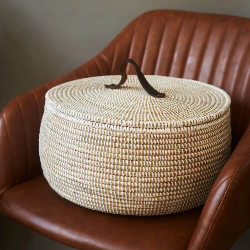 The image shows a large woven basket made in Senegal, sitting on a brown leather chair. The basket is round and has a lid, with a brown leather handle attached to the top. The basket is woven with alternating stripes of white and tan, creating a textured pattern. The lid fits snugly on top of the basket, and the handle is positioned in the center of the lid. The chair is made of brown leather and has a smooth, glossy finish. The chair has vertical ridges. The surface of the chair is smooth and slightly reflective. The background is a neutral color, which helps to highlight the basket and chair. The lighting is soft and diffused, which creates a warm and inviting atmosphere. The basket appears well-made and sturdy. The leather handle looks strong and durable.