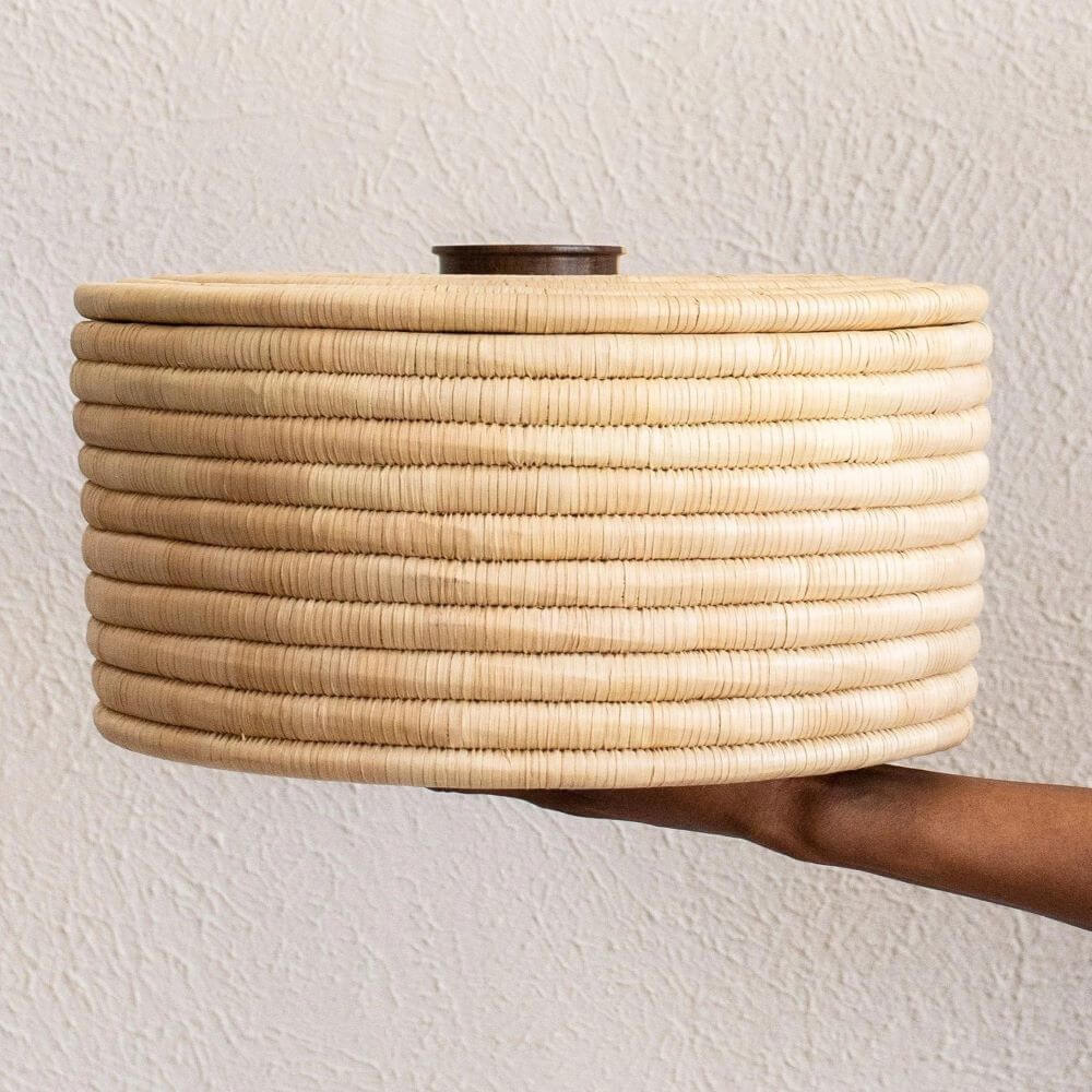 The photograph presents a close-up view of a round, lidded basket being held by a dark-skinned hand. The background is a textured, off-white wall with a subtle, mottled pattern. The basket is composed of a light beige, woven material that appears like straw or raffia, meticulously coiled to form a sturdy, horizontal-striped pattern. The individual coils are tightly wound, creating a ridged texture. A circular lid covers the basket, crafted from the same woven material, topped with a dark brown, cylindrical wooden knob for easy lifting. A brown arm and hand provide support beneath the basket. The focus is sharp on the basket and the hand holding it, with a slight depth of field blurring the wall in the background. The lighting is soft and even, casting gentle shadows that accentuate the texture of both the basket and the wall. The image is well-composed, highlighting craftsmanship and materials used in making the basket.