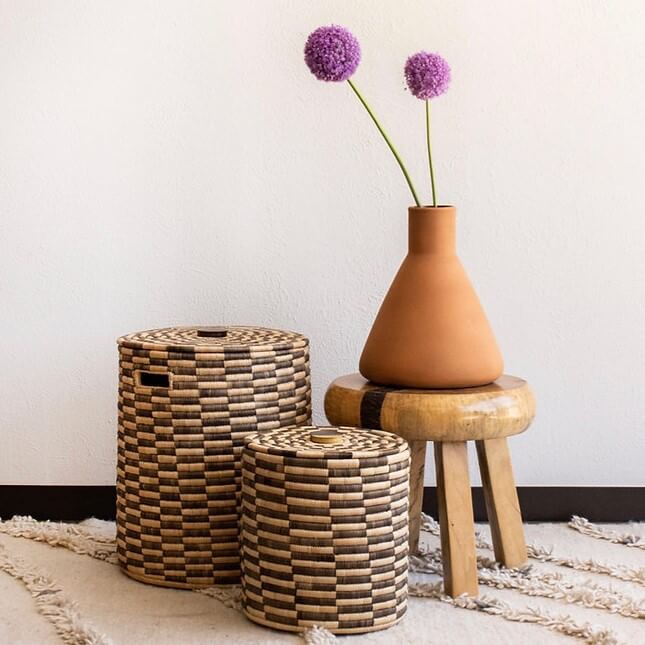 The image showcases a minimalist indoor scene. The backdrop is a plain white wall, against which a still life arrangement is composed. In the foreground, there are two cylindrical African baskets with lids, arranged at slightly different depths to create perspective. The baskets feature a woven pattern of alternating light and dark brown hues, giving them a textured appearance. To the right of the baskets, a small wooden stool stands. It is a rustic piece with a round top and three wooden legs. A brown, terracotta vase sits on the stool, holding two vibrant purple allium flowers. The long green stems of the flowers reach upwards, adding a touch of natural color to the setting. The entire arrangement is placed on a cream-colored rug with a shaggy texture. This adds depth and softness to the composition. The light in the image is soft and diffused, creating a warm and inviting atmosphere.