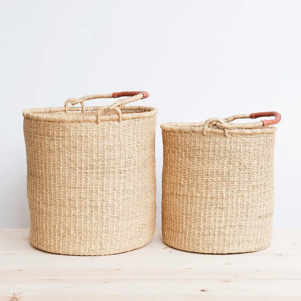 The image shows two woven laundry African baskets of different sizes, positioned side by side on a light wooden surface. The backdrop is a plain white wall, providing a clean and neutral background. Both baskets are cylindrical and appear to be handwoven from a natural-colored, straw-like material. The weaving is intricate, with a tight pattern that gives the baskets structure and visual texture. The larger basket is on the left and is taller and wider than the one on the right. It has a pair of sturdy handles made from a similar woven material, reinforced with what appears to be a reddish-brown leather grip at the center of each handle. The handles are attached to the rim of the basket on opposite sides, allowing for easy carrying. The smaller basket mimics the design of its larger counterpart but is proportionally smaller. It also has woven handles with leather grips. The baskets are placed on a light-colored wooden surface, which subtly contrasts with the neutral tones of the baskets. The wood grain is visible, adding a touch of natural texture to the image. The lighting is soft and even, casting gentle shadows that highlight the woven texture of the baskets and their cylindrical forms.