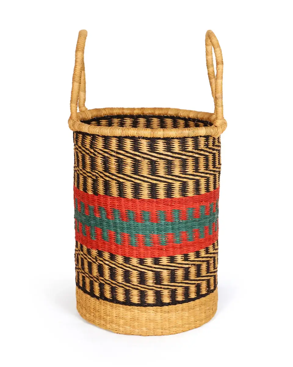 A handwoven laundry basket is prominently displayed against a stark white backdrop, its cylindrical form emphasized by a subtle shadow cast on the surface beneath. Made from natural straw, the basket showcases horizontal bands of alternating light and dark brown shades, creating a rhythmic pattern that envelops its entire body. A broader band of solid brown with dark accents is placed around the center of the basket. This highlights a recurring geometric pattern. The dark and turquoise accents are created by woven details that appear like short, bold dashes in rows. The basket’s sturdy construction extends to its rim, which is reinforced with a thicker band of straw, ensuring structural integrity and durability. Two cylindrical handles, also crafted from straw, stand upright from the rim, providing a comfortable grip for carrying. The handwoven texture is readily noticeable across the basket's surface. It adds to the basket's aesthetic appeal and attests to its handmade nature. The consistent weave pattern and the basket’s well-preserved condition attest to its quality craftsmanship.