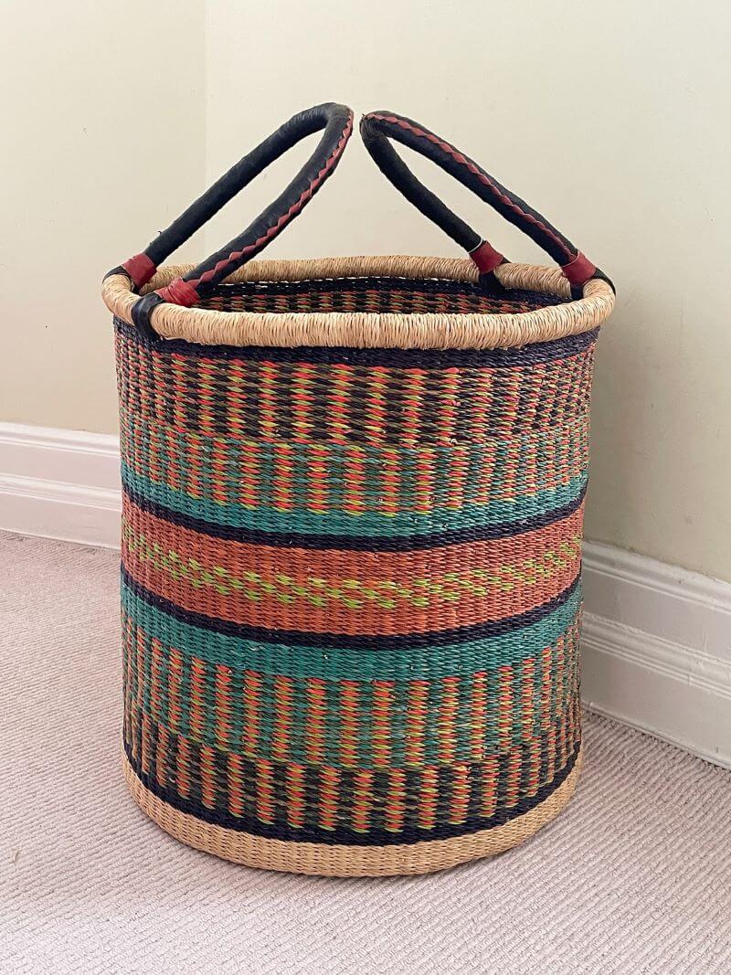 The image shows a hand-woven laundry basket indoors. The basket's cylindrical body begins with a base and gradually extends upwards. The primary material of the basket appears to be straw, meticulously woven in a close-knit pattern. The design stands out with alternating bands of intricately woven colors. The colors are arranged with precision, creating a visually appealing harmony. The topmost part of the basket is completed with a straw border, which is finished off with a darker strip secured by two handles on opposite sides. The handles are notably thicker, sheathed in black material bordered in red stitching along the sides. The basket sits on a neutral-colored carpet, up against a beige wall detailed with white baseboards. The lighting in the room is soft and even, accentuating the various textures of the carpet, wall, and woven surface. The basket's design and structure evoke a sense of traditional craftsmanship, while its coloring gives it a contemporary aesthetic look.