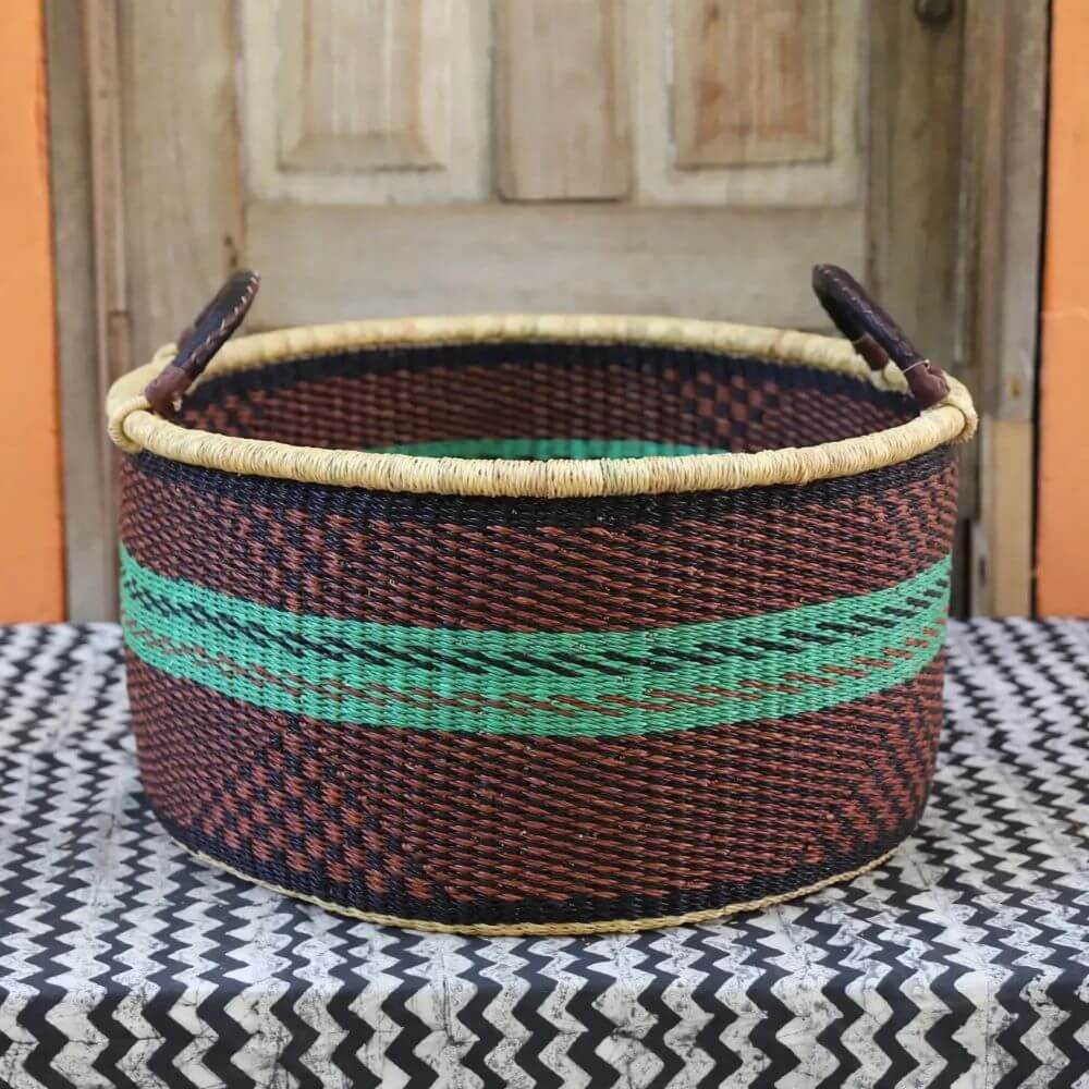 This image displays a handwoven basket with a traditional aesthetic, positioned on a table with a distinctive zigzag pattern. The basket is the image's focal point, showcasing its round shape and intricate woven texture. The primary colors of the basket are a dark brown and black, contrasted by a horizontal band of turquoise that encircles the midsection. The rim of the basket is adorned with a lighter, natural-colored trim which gives a neat and structured finish. There are also two handles made from similar woven material, attached to the upper part of the basket with leather accents, rendering it easy to carry. The table upon which the basket sits is covered with a black and white zigzag patterned cloth that adds a sense of depth and contrast to the image. In the background, there is a rustic wooden door with neutral tones that complements the basket’s earthy colors and textures. A glimpse of an orange surface adjacent to the door provides a subtle pop of color, enhancing the overall composition. The lighting is natural, casting soft shadows that highlight the basket's texture and form. The composition is well-balanced, with the basket taking center stage and the background elements providing context without overpowering the main subject. The image is likely intended to showcase the craftsmanship and aesthetic appeal of the handwoven basket, potentially for marketing or artistic purposes.