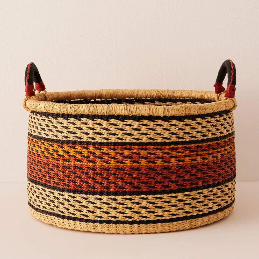 The image showcases a cylindrical woven basket with two looped handles, set against a plain, off-white backdrop. Popular storage shape among African baskets. The basket's body is primarily a warm, golden straw color, interwoven with black to create patterned bands. The pattern consists of rows where pairs of straw-colored stitches alternate with pairs of black stitches, adding a touch of geometric flair to the texture. Around the center of the basket, a wide band displays a gradient of vibrant orange and deep reddish-brown hues, which are likely from the dyes used on the material of the basket. Flanking these colors are two single bands of solid black, outlining the center pattern and adding to their contrast. The lip of the basket is framed with a thicker, wound material that is a blend of natural straw tones, maintaining the earthy palette of the rest of the container. Two looped handles are attached to the basket’s rim with red and black leather. The handles are angled outward and positioned in such a way that suggests they are functional for lifting and moving the basket. The even lighting casts soft shadows, which helps to define the weaving without overly emphasizing individual strands, producing a clear distinction in material colors and construction.