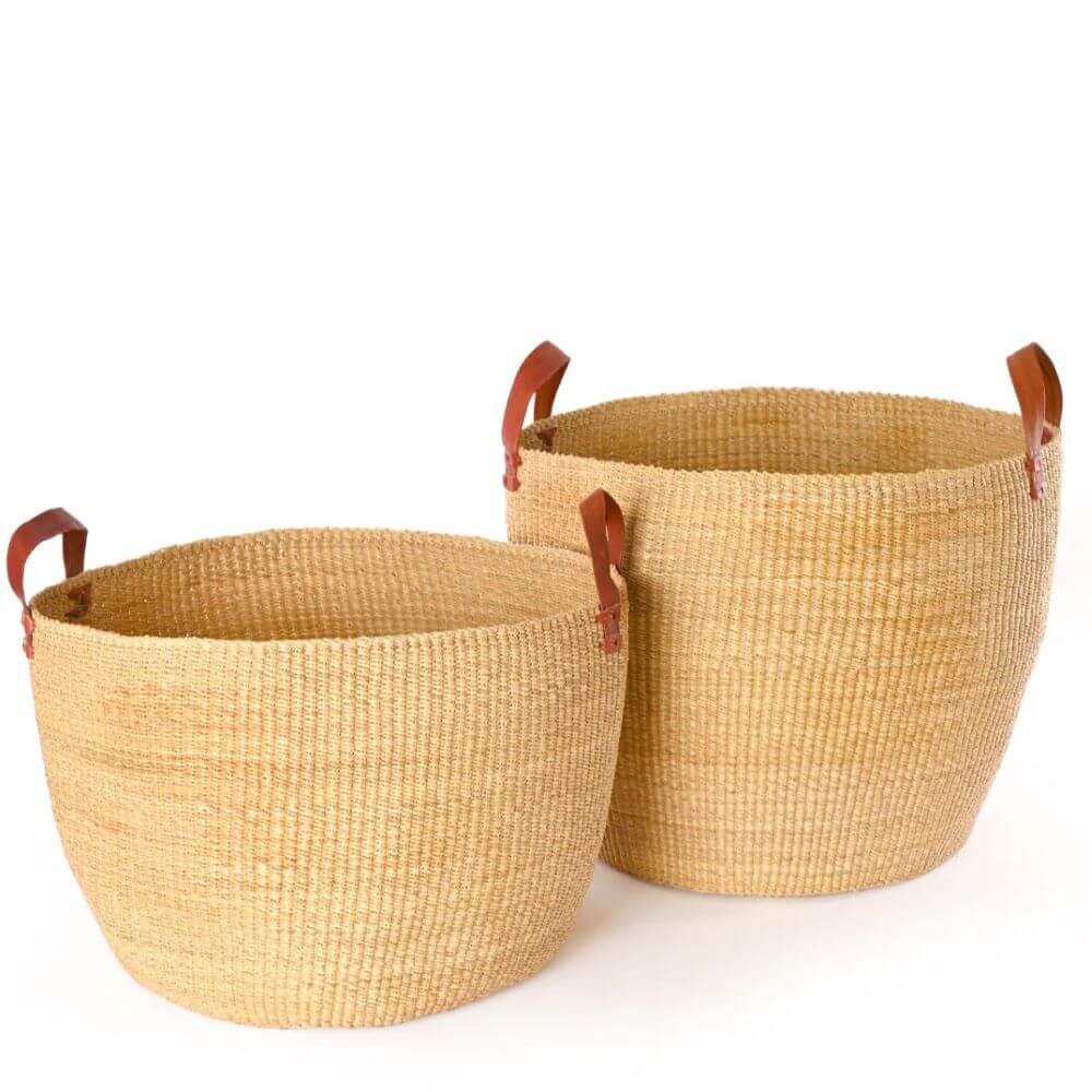 The image shows two light tan woven African baskets of varying sizes, positioned next to each other against a plain white background. The basket on the left is smaller and has a wider, more open shape. The basket on the right is taller and more cylindrical. Both baskets are crafted from a tightly woven material that gives them a textured, ribbed appearance. Each basket features handles made of brown leather, attached to the sides with fasteners, adding a touch of sophistication and practicality to the design. The texture of the woven material provides a rustic contrast to the smooth leather handles. The lighting in the image is bright and even, highlighting the natural tones and texture of the baskets. The plain white background helps to keep the focus on the baskets, showcasing their form and material. The style and detailing of the baskets suggest that they are handcrafted.