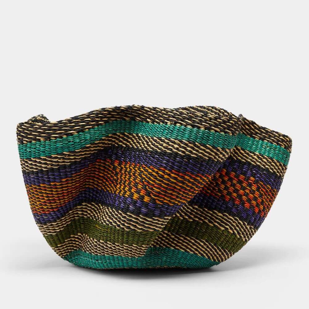 A close-up studio shot showcases a round, woven pakurigo wave basket featuring a dense, banded pattern against a plain white backdrop. The basket is predominantly composed of tightly intertwined natural fibers, with alternating bands of color enriching its form. The fiber color palette consists of alternating black and sand-colored strands. Prominent horizontal bands in various colors punctuate the woven texture. These include shades of green, orange, and purple hues that add to the basket's vibrant mosaic. The weaving is intricate and uniform, providing a tactile sense of the basket's handmade nature. The basket stands firmly on its base, with the sides curving inward slightly. The overall effect is one of organic craftsmanship melded with an understated elegance.