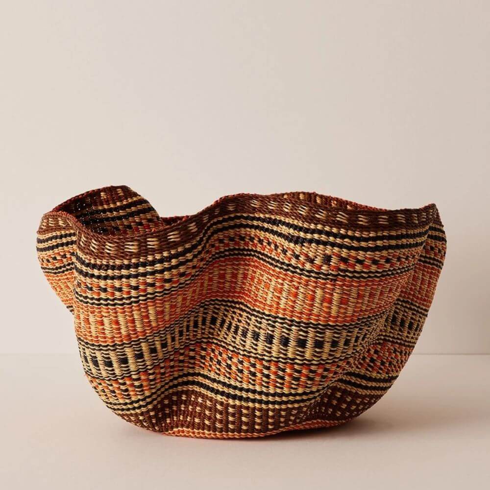 This image features a large woven pakurigo wave basket with a distinctive wavy upper edge. The basket is made from natural fibers, exhibiting a mixed pattern in shades of brown, black, and bright orange. The weaving is intricate, arranged in horizontal stripes that vary in width and pattern. Some stripes are simple bands of solid color, while others consist of repeating geometric elements such as thin dashes and small squares, creating a textured visual effect. The overall shape of the basket is rounded at the bottom and expands upwards, culminating in an irregular, wavy rim. This uneven edge gives the basket a natural, handcrafted appearance. The basket sits on a neutral surface, likely a tabletop or a similar flat area, against a plain, light-colored backdrop. The lighting in the image is soft and diffused, which highlights the texture and colors of the woven material without creating harsh shadows. The neutral setting allows the basket to be the sole focus, emphasizing its unique shape, pattern, and craftsmanship.