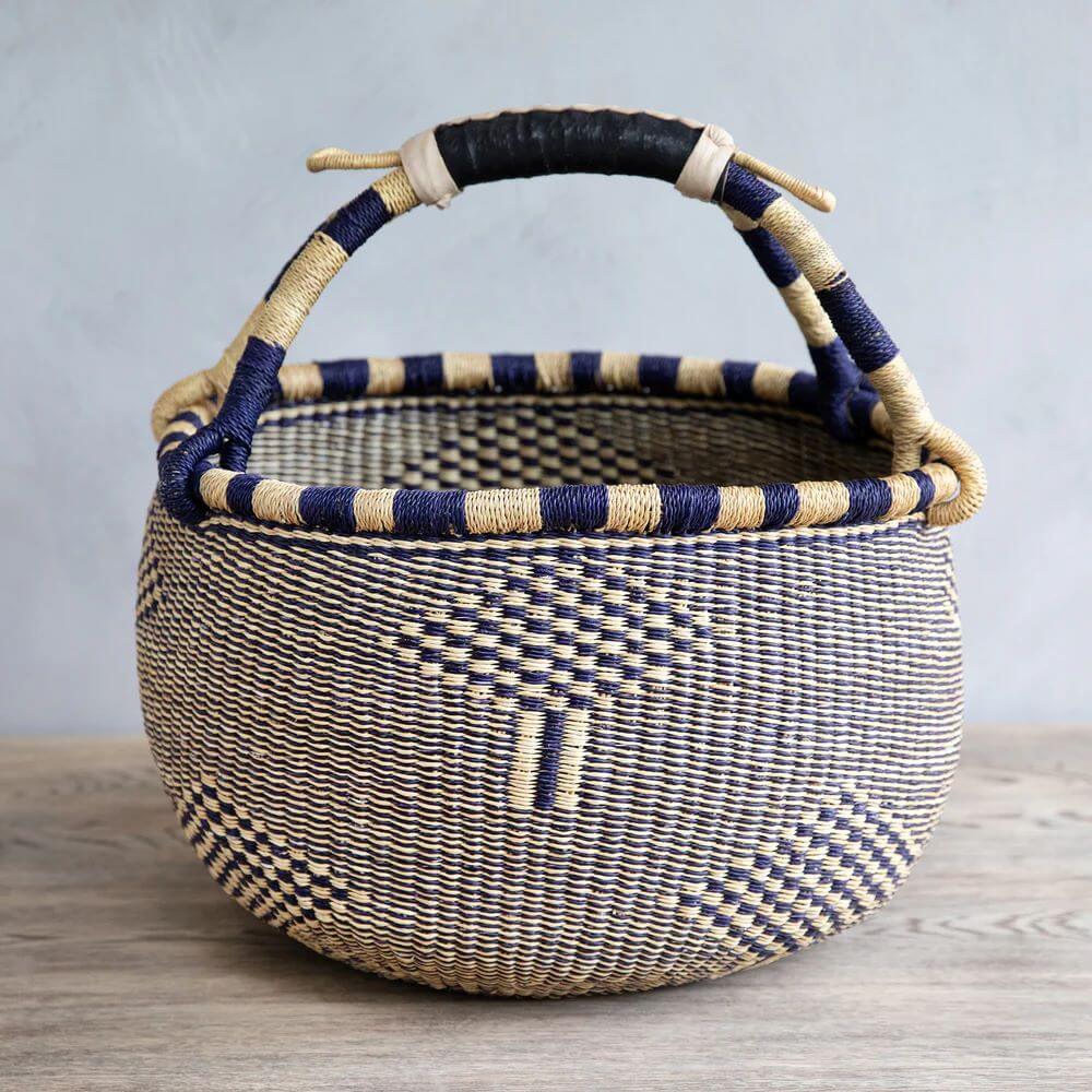 The image shows a traditional handwoven bolga basket, shape from Ghana popular among African baskets, with a distinctive design and construction. The basket is primarily cylindrical, with its form slightly bulging outwards from the base before curving inward towards the brim. The base is stable and the basket stands upright on a light wooden surface. Crafted from natural fibers, the basket displays a woven checkerboard pattern. This pattern alternates between a dark navy blue and a light tan color, which creates a textured, dimensional appearance. The exterior of the basket body is predominantly covered with this pattern but also includes other design elements. Notably, there are vertical stripes of the tan color, providing visual interruption and subtle detailing. Along the rim of the basket, a band is woven with alternating dark blue and tan stripes, which adds a refined finish to the basket's edge. The rim is not perfectly straight but rather has slight variations in height, indicating its handmade nature. A sturdy handle extends from the rim, connected to the basket on both sides. The handle is largely wrapped in tan-colored fiber, matching the overall color scheme. At the apex of the handle, there's a section wrapped in black material, possibly leather or a similar durable substance. The ends of the fiber wrapping are secured in a rudimentary manner, with loose ends sticking out. The background is nondescript, featuring a smooth, gray wall that contrasts with the basket’s texture and color. This helps to emphasize the basket as the primary subject of the image. In summary, the basket is a handcrafted item with an emphasis on geometric design and traditional weaving techniques. The use of natural materials and its handmade quality are clearly evident.