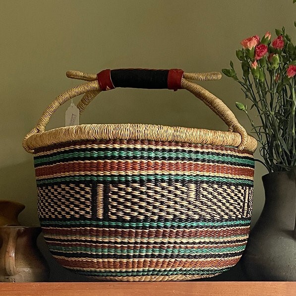 An eye-level, medium shot shows a woven basket in the foreground to the left of a dark grey vase with pink carnations against an olive green wall. A section of what looks like a wooden surface forms the bottom one-fifth of the image. The basket takes up the center of the image. Below the basket’s light natural top border is a colorful woven pattern that includes bands of brown, light blue and black, with a block of black and tan diamond shapes in the center. The natural color handles are covered with black and red leather in the center. A tag is attached to one of the handles. To the left of the basket in the foreground is a piece of a light brown pot or vase that is cut off. The dark grey vase takes up the right edge of the image and has pink carnations in it. The flowers are in full bloom and some buds have yet to open. The olive green wall serves as the background.