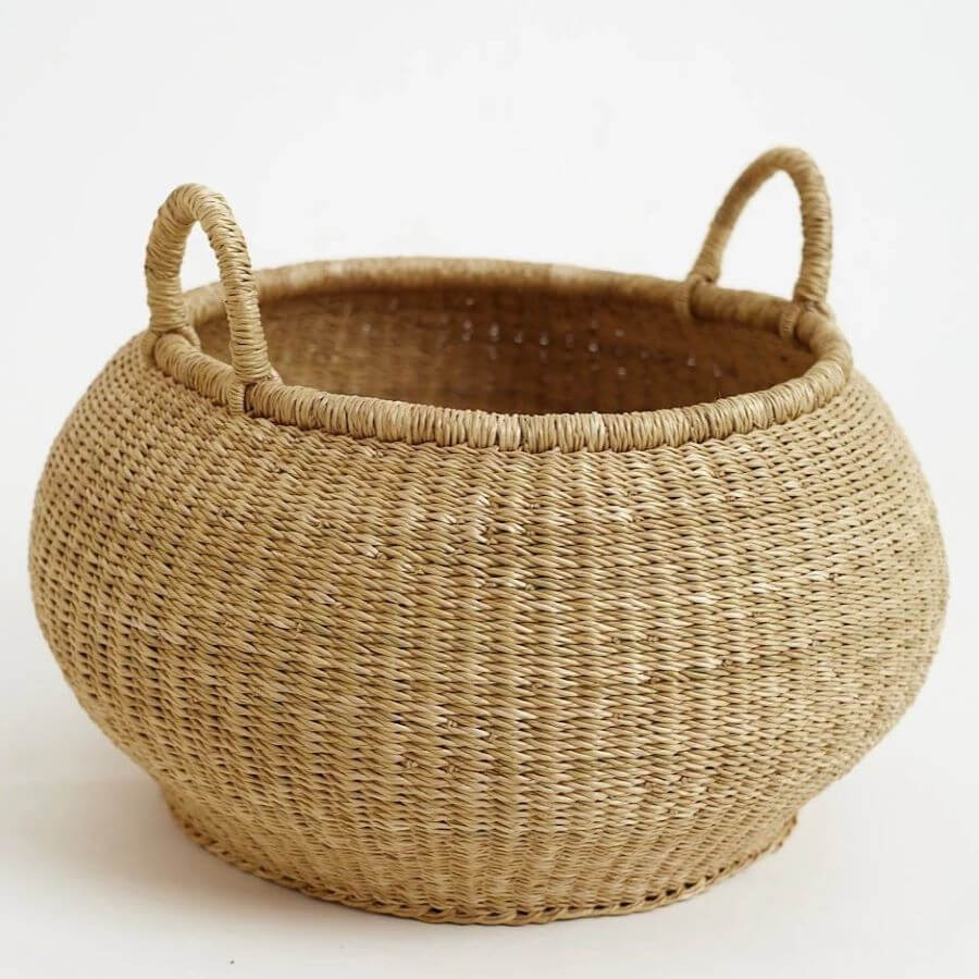 The image showcases a handwoven, round bolga storage basket with handles, set against a clean, white background. The basket's textured surface and natural color give it a rustic and organic appeal. Basket Details: Shape: The basket features a rounded, almost spherical body, narrowing slightly towards the base for stability. Material and Texture: The basket is constructed from woven natural fibers (likely straw or a similar material), creating a textured, tactile surface with small variations in color and weave. Color: The basket is predominantly a light beige or light brown color Handles: Two sturdy handles are attached to the upper part of the basket. Weave: The weaving pattern appears consistent and tight, providing strength and durability to the basket. Rim: The basket has a finished rim, likely reinforced with additional weaving or a different material to maintain shape and prevent fraying. Background: Color: The backdrop is a bright, clean white, providing a high contrast that highlights the basket. Surface: The surface appears smooth and uniform, avoiding any distractions from the product. Overall Composition: The image is well-lit and focuses squarely on the basket, providing a clear and detailed view. The simplicity of the background and setting accentuates the basket's natural aesthetic.