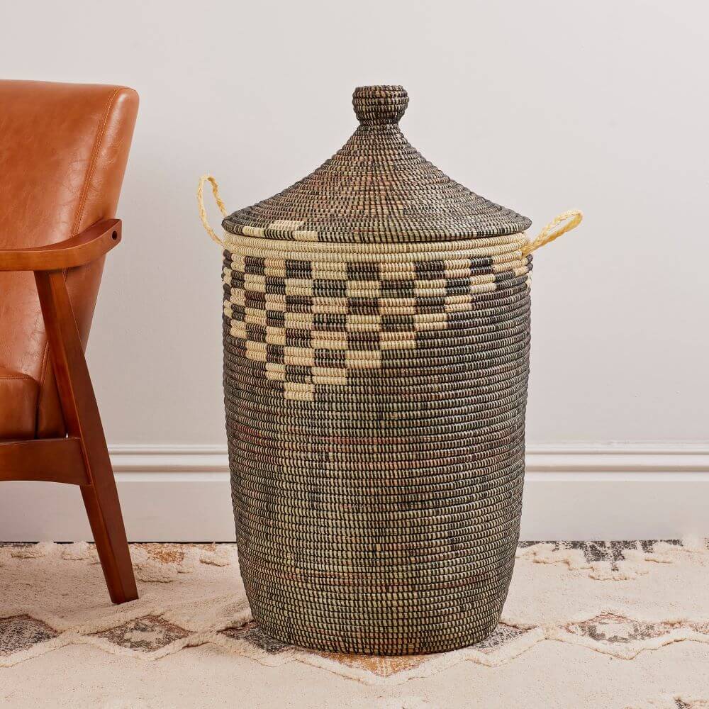 The image shows a tall, cylindrical woven Senegalese hamper basket with a lid, situated on a patterned rug next to a mid-century style arm chair. The basket itself is intricately woven, composed of dark brown or black and a beige or natural colored wicker. The cylindrical body rises smoothly before transitioning into a decorative band of alternating squares, forming a checkerboard pattern around the upper part of the basket. At the top, the basket is capped with a conical lid that has a small, round knob at its peak. Two small handles made of rolled wicker protrude from the sides of the basket. The rug beneath is patterned with geometric shapes and subtle colors. Next to the basket, on the left, is the arm of a high-back chair. The chair is made of a brown leather with a dark brown wooden frame. The background is plain, with a hint of an off-white wall behind the basket and chair, allowing the intricate details of the basket and rug to stand out. The lighting appears soft and natural, highlighting the textures and colors of the woven materials.