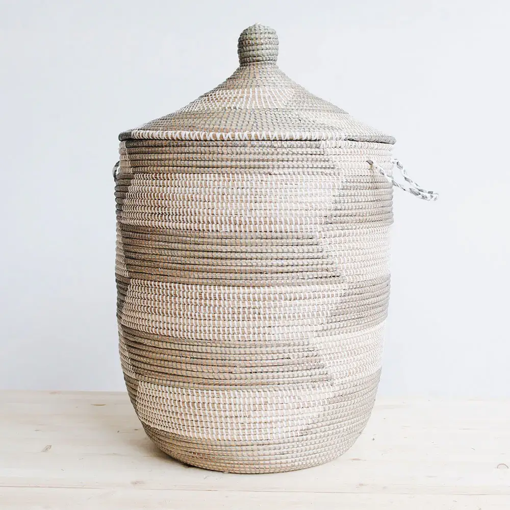 The image features a tall, cylindrical Senegalese hamper basket with a lid, showcasing a woven design in alternating gray and off-white tones. The basket is placed on what appears to be a light wooden surface against a white background, emphasizing its form and texture. The basket has a rounded base that gradually widens before maintaining a consistent cylindrical shape through most of its height. Horizontal bands of alternating gray and off-white weave encircle the basket, creating a striped pattern that is rhythmic and visually appealing. The weave appears tight and uniform, indicating careful craftsmanship. Small handles are visible on the sides, suggesting the basket is functional and meant to be moved. The lid of the basket is conical and tapers to a rounded knob at the top. Its woven pattern mimics that of the basket, with alternating bands that seamlessly integrate into the overall design. The lid sits snugly on the basket, completing the enclosure and adding to the piece's aesthetic appeal. The lighting in the image is soft and diffused, highlighting the texture of the woven material and casting gentle shadows that define the basket's shape. The color palette is neutral, with the gray and off-white tones creating a sense of calm and simplicity, while ensuring the basket remains the central focal point.