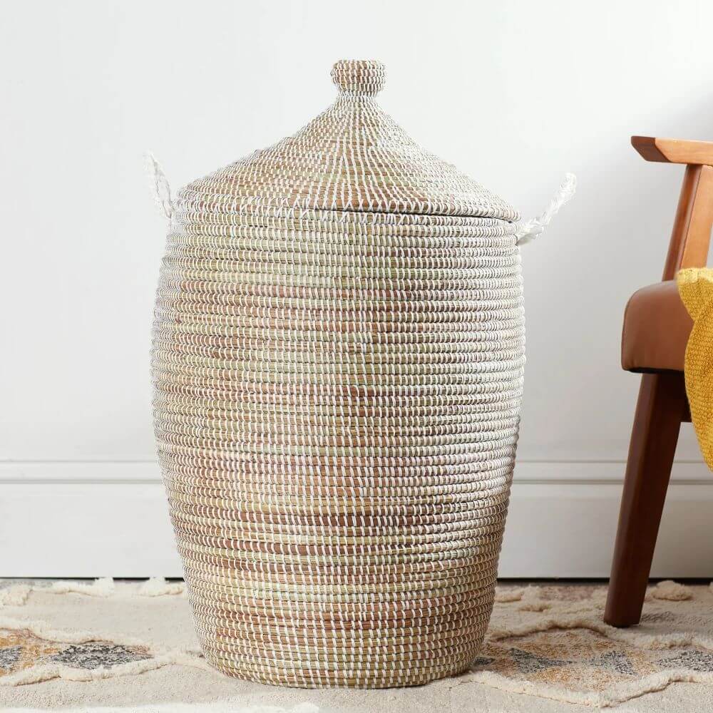 A tall, cylindrical Senegalese hamper basket with a lid stands prominently next to a chair, set against a minimally decorated backdrop. The basket's texture is defined by its weave of light, natural fibers interspersed with white strands that create horizontal lines. The color of the fibers varies from light tan to darker browns, contributing to its rustic, handcrafted appearance. The basket gradually narrows towards the top, where it is crowned with a conical lid. This lid features a small, round knob at its peak, serving as a handle. Two rope handles are attached to the basket’s sides, adding a practical element for carrying or moving it. To the right of the basket, a mid-century modern chair is positioned with its dark wooden leg visible. Part of its brown, leather-upholstered seat and a swatch of yellow fabric draped over the chair add warmth to the scene. Beneath the basket and chair, the floor is covered with a plush, patterned rug. The rug's design includes subtle geometric shapes and earthy tones, enhancing the overall aesthetic. The backdrop consists of a plain white wall with a simple baseboard, providing a clean and neutral setting that allows the basket and chair to stand out. The lighting is soft and diffused, casting gentle shadows and highlighting the textures and colors of the objects. The setting suggests a well-curated interior space, possibly a living room or bedroom, emphasizing the basket's functionality and decorative appeal.