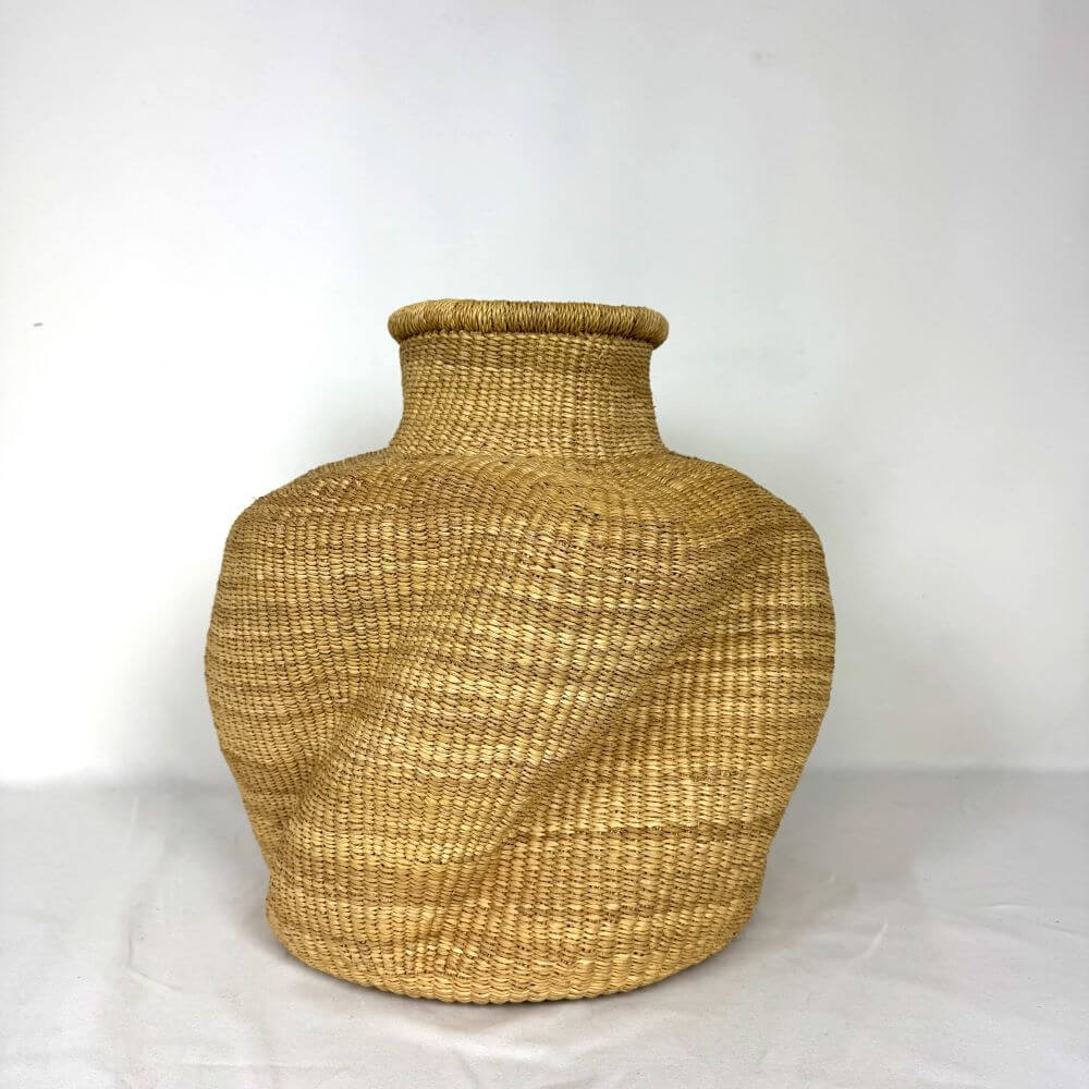 The image shows a handmade Bolga pot basket, a traditional basket made by artisans in the Bolgatanga region of Ghana. These baskets are known for their durability and unique designs. The pot has a distinctive form. The body tapers upwards from the base to a rounded midsection, and then gently curves inward to form a narrow neck. The lip of the pot is reinforced with a rolled edge that gives it a clean, finished appearance. There is a diagonal weave design in the center of the pot and a vertical weave design above and below the central design. The overall color of the basket is a golden tan, with subtle variations in tone that highlight the natural shades of the straw. The basket is placed on a white surface, which helps to accentuate its shape and texture. The neutral background helps to maintain focus on the pot itself, rather than distracting from the details.