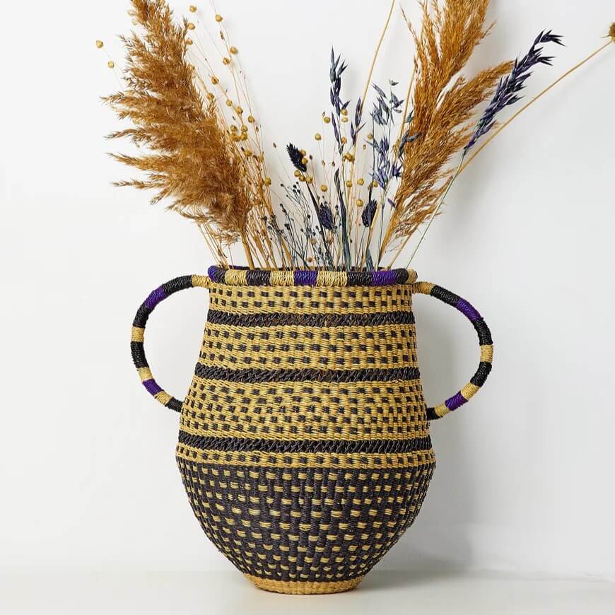 This eye-level, full, and medium shot showcases a woven vase basket brimming with dried botanical elements. The vase, a blend of tan and black, exhibits a traditional weaving pattern throughout. Its base is woven entirely in black with tan undertones, transitioning into layers of horizontal stripes. Wider black bands are interspersed between finer tan and black weaving, which makes up the majority of the structure. The lip of the vase is embellished with a decorative black-and-tan checkerboard pattern, bordered by vibrant purple accents. The handles of the vase curve outward and are partly colored in black with stripes of tan and purple. The vase is filled with assorted botanical elements, a mix of light brown pampas grass, delicate wildflower stems, and some darker purple stems. The backdrop is a plain white wall and the base of the vase rests securely on a flat, off-white surface. The overall composition is clean and well-lit, accentuating the intricate details of the woven vase and the rustic beauty of the dried plants.