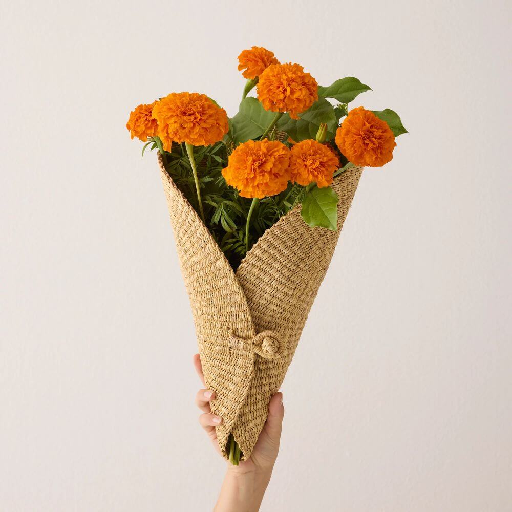 A hand holds a bouquet of orange marigolds, wrapped in a tan woven flower cone. The marigolds, with their pom-pom-like blossoms, are in full bloom, their vibrant orange contrasting against the green stems and leaves. The woven bag is made of a natural fiber and has a rough texture. The bag is folded and wrapped around the flowers in a cone shape, secured with a knot at the middle. The hand holding the bouquet is pale and the fingers are gently curved around the base of the bag. The background is a plain, off-white wall, which provides a clean backdrop that allows the colors and textures to be the primary focus. The lighting is soft and diffused, giving the image a gentle, natural feel. The composition is simple, with the bouquet positioned in the center of the frame.