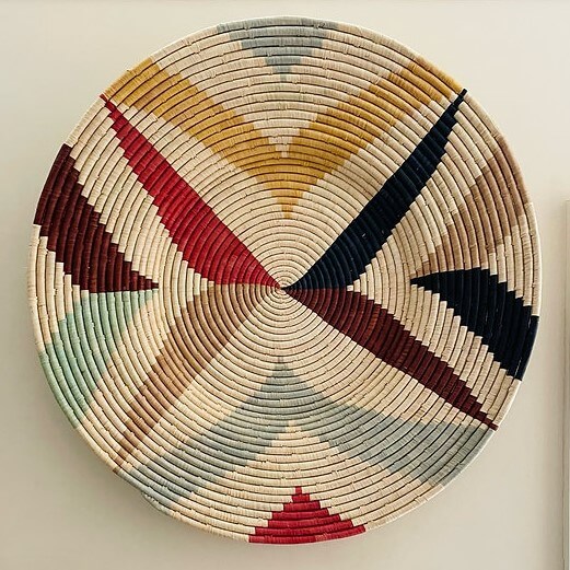 The image features a decorative woven wall basket or shallow basket, typical of African crafts, mounted on a wall, likely for display. The plate has a circular shape and is made from natural fibers, appearing to be tightly coiled and sewn together to form concentric circles. The natural color of the fibers serves as the base, a light beige or ecru, creating a neutral backdrop. There are triangular patterns created with various colored fibers, resembling rays emanating from the center. The colors include shades of yellow, red, navy blue, light blue, and burgundy. These colors form distinct, bold patterns that contrast against the natural fiber background. The weaving technique is evident in the texture, with the individual strands of fiber visible, creating a tactile appearance. The concentric circle pattern suggests a coiled basketry technique, where a continuous coil of fiber is spiraled and stitched together as it grows outward. The plate is positioned against a plain, creamy white wall, which provides a simple and uncluttered backdrop that highlights the intricate design and colors of the woven piece. The lighting is even, casting minimal shadows and allowing the artwork to be viewed clearly and without distortion. Overall, the image is a sharp, well-lit representation of an African art piece used for decorative purposes.